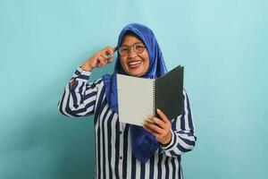 opgewonden Aziatisch middelbare leeftijd vrouw, vervelend een blauw hijaab, bril, en een gestreept shirt, houdt een boek en past zich aan haar bril terwijl op zoek direct Bij de camera, staand tegen een blauw achtergrond. foto