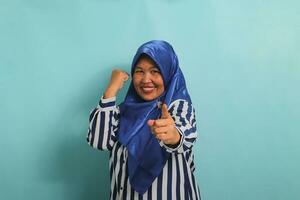 een opgewonden middelbare leeftijd Aziatisch vrouw in een blauw hijab en gestreept overhemd shows een duimen omhoog en een Oke teken, symboliseert succes en uitdrukken goedkeuring, terwijl staand tegen een blauw achtergrond. foto