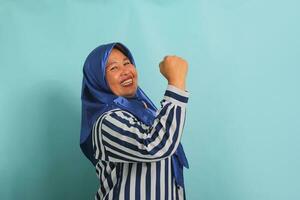 een enthousiast middelbare leeftijd Aziatisch vrouw, in een blauw hijab en een gestreept shirt, is gezegde Ja met een gelukkig uitdrukking, vieren zege met een vuist pomp gebaar terwijl staand over- een blauw achtergrond foto