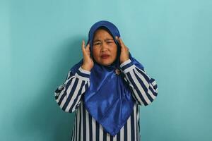 portret van een middelbare leeftijd Aziatisch vrouw, vervelend een blauw hijab en gestreept shirt, shows haar lijden van een hoofdpijn, migraine, of ziekte, staand tegen een blauw achtergrond foto