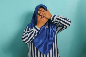 een ongelukkig middelbare leeftijd Aziatisch vrouw in een blauw hijab is aan het bedekken haar gezicht en aanraken haar voorhoofd, uitdrukken droefheid, depressie, en angst, terwijl staand tegen een blauw achtergrond foto