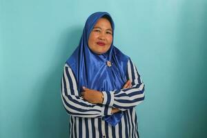 een glimlachen middelbare leeftijd Aziatisch vrouw, vervelend een blauw hijab en een gestreept shirt, staat met haar armen gekruist, uitstralend geluk en vertrouwen tegen een blauw achtergrond foto