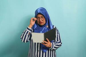 nadenkend Aziatisch middelbare leeftijd vrouw, vervelend een blauw hijaab, bril, en een gestreept shirt, houdt een Open boek en accenten haar bril met een onzeker uitdrukking terwijl staand tegen een blauw achtergrond foto