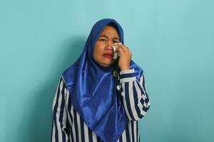 een van streek middelbare leeftijd Aziatisch vrouw, vervelend een blauw hijab en een gestreept shirt, is huilen en afvegen tranen met een zakdoek, uitdrukken frustratie en nood terwijl staand tegen een blauw achtergrond foto