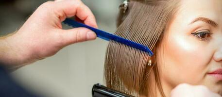 kapper rechtzetten vrouw bruin haar- van medium lengte met een haar- met een ijzer haar- stijltang en kam in een schoonheid salon. foto