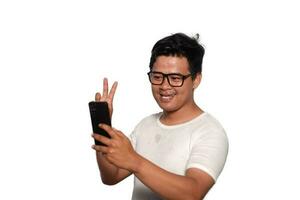 opgewonden Aziatisch Mens met bril vervelend wit t-shirt glimlachen terwijl Holding zijn telefoon, geïsoleerd door wit achtergrond foto