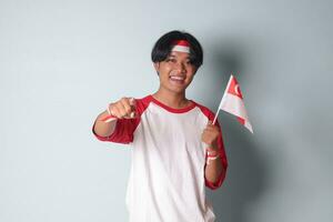 portret van aantrekkelijk Aziatisch Mens in t-shirt met rood en wit lint Aan hoofd, tonen Product en aanwijzen weg terwijl Holding Singapore vlag. geïsoleerd beeld Aan grijs achtergrond foto