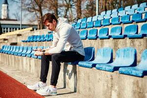 Mens in sportkleding resting Bij stadion stoel en gebruik smartphone foto