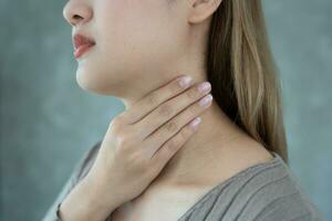 Aziatisch vrouw zetten haar hand- Aan haar nek ten gevolge naar een zeer keel. vrouw tonsillitis oorzaken een ontsteking. behandeld door nemen water, nemen antibiotica, raadplegen een dokter, virus, corona virus, influenza, griep foto