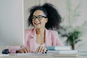 gekruld vrouw manager glimlacht, werken van een afstand in elegant pak, bril, Bij bureaublad. foto