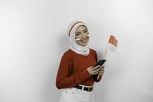 een glimlachen Aziatisch moslim vrouw vervelend rood top en wit hijaab, Holding haar telefoon en vlag, geïsoleerd door wit achtergrond. Indonesië's onafhankelijkheid dag concept foto