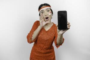 jong mooi vrouw vervelend een rood kebaya is tonen haar telefoon terwijl geschreeuw en schreeuwen luid met een hand- Aan haar mond. Indonesië's onafhankelijkheid dag concept. foto