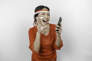 jong mooi vrouw vervelend een rood kebaya is Holding haar telefoon terwijl geschreeuw en schreeuwen luid met een hand- Aan haar mond. Indonesië's onafhankelijkheid dag concept. foto