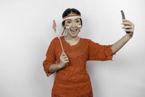 een gelukkig Aziatisch vrouw vervelend rood kebaya en hoofdband, Holding haar telefoon en Indonesië's vlag, geïsoleerd door wit achtergrond. Indonesië's onafhankelijkheid dag foto