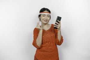 een gelukkig Aziatisch vrouw vervelend rood kebaya en hoofdband, Holding haar telefoon, en gebaren liefde teken door haar vingers tonen inschrijving gevoel, geïsoleerd door wit achtergrond. Indonesië's onafhankelijkheid dag foto