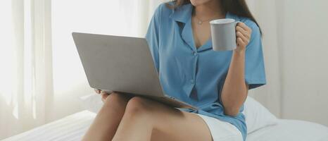 jong Aziatisch vrouw met mooi gezicht, lang haar, blauw overhemd aan het liegen Aan bed in wit slaapkamer Bij huis met kop van koffie en video telefoontje met laptop pratend naar familieleden Aan vakantie. vakantie concept. foto