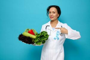 mooi vrouw, vrouw dokter voedingsdeskundige vervelend een wit medisch japon met blauw bewustzijn lint points Aan een bord vol van gezond rauw veganistisch aan het eten. wereld diabetes dag concept met ruimte voor advertentie foto