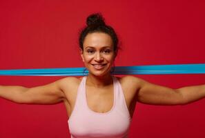 detailopname van een charmant vastbesloten sportvrouw oefenen met elastisch geschiktheid band, uitrekken haar armen tegen rood achtergrond met kopiëren advertentie ruimte foto