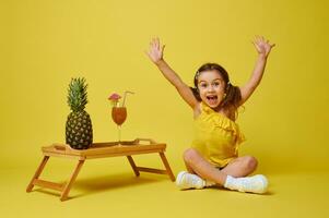 aanbiddelijk weinig meisje verhogen haar armen omhoog en uitdrukken geluk terwijl zittend Aan geel achtergrond in de buurt een portie bamboe dienblad met een ananas en cocktail. zomer concept foto
