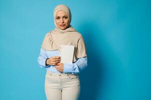 Arabisch moslim vrouw met een wit boek in haar handen. onderwijs concepten Aan blauw achtergrond met kopiëren ruimte foto