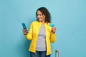 Afrikaanse Amerikaans mooi vrouw vervelend geel jasje, glimlacht Holding blauw credit of korting kaart met kopiëren ruimte en vegen, browsen websites, boeken Aan mobiel telefoon, geïsoleerd over- blauw achtergrond foto