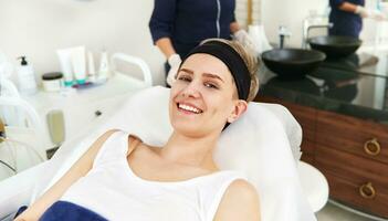 mooi vrouw met mooi glimlach ontspannende Aan massage tafel Aan de achtergrond van wazig onherkenbaar dokter schoonheidsspecialist dermatoloog foto