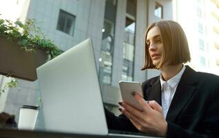 zelfverzekerd gemotiveerd jong freelance, bedrijf vrouw gebruik makend van mobiel telefoon en werken Aan laptop zittend buitenshuis Aan stedelijk achtergrond. effectiviteit, online bedrijf concept foto