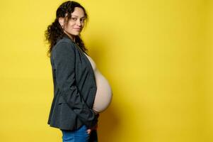 mooi zwanger vrouw in zwangerschap 36 week, glimlacht Bij camera, voelt gelukkig emoties ervan uitgaand baby, geel achtergrond foto