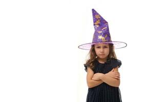 portret van schattig weinig heks boos van streek meisje vervelend een tovenaar hoed en gekleed in elegant carnaval jurk, op zoek Bij camera poseren met gekruiste armen tegen wit achtergrond, kopiëren ruimte, halloween foto