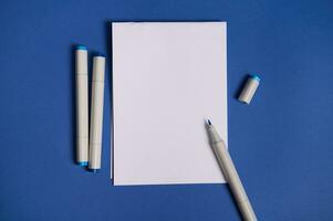tweekleurig vlak leggen samenstelling met waterverf markeringen of viltstift pennen met een wit blanco leeg papier vel met kopiëren ruimte, geïsoleerd Aan blauw achtergrond. foto