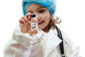 mooi weinig meisje in dokter uniform op zoek Bij vaccin flacon Aan haar hand. geïsoleerd Aan wit achtergrond met kopiëren ruimte foto