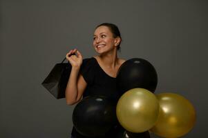 zwart vrijdag concept met een vrolijk vrouw met mooi toothy glimlach, gekleed in zwart Holding zwart en gouden lucht ballonnen en boodschappen doen tas, op zoek de kant Bij een grijs achtergrond, kopiëren advertentie ruimte foto