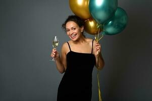 Afrikaanse Amerikaans mooi vrouw met glimmend gouden groen metalen lucht ballonnen poses tegen grijs muur achtergrond met kopiëren ruimte, looks Bij camera Holding een glas van sprankelend wijn in handen. foto