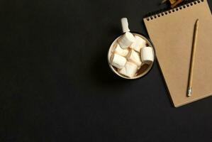 vlak leggen samenstelling met houten potlood Aan ambacht kladblok met leeg blanco vel en een kop van heet cacao drinken met heerlijk zoet wit marshmallows Aan zwart achtergrond, kopiëren ruimte. Kerstmis concept foto