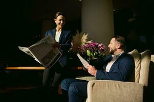 twee knap mensen, midden- oud Kaukasisch Mens en mooi elegant vrouw in bedrijf pak, collega's, bedrijf partners Aan bedrijf reis, pratend en lezing een krant- in de hotel lobby foto