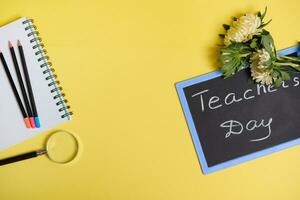 vlak leggen van asters bloemen aan het liegen naar beneden Aan schoolbord met tekst leraren dag , en een vergrootglas loupe De volgende naar gekleurde potloden Aan een blanco lakens van een kladblok geïsoleerd Aan geel achtergrond met kopiëren ruimte foto