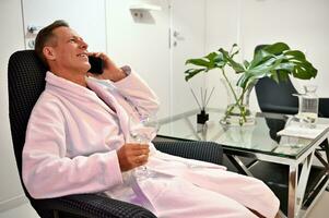 ontspannen knap midden- oud 50 jaren oud Mens in wit badstof badjas zittend Aan een fauteuil met een glas van vers water en pratend Aan mobiel telefoon Bij welzijn spa centrum foto