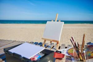 blanco wit canvas Aan een houten tafel ezel De volgende naar verven en borstels Aan een houten dek stoel Aan de strand tegen de achtergrond van mooi blauw zee foto