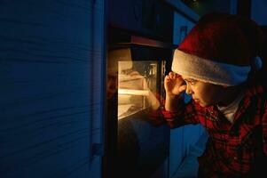 aanbiddelijk knap nieuwsgierig Kaukasisch jongen in de kerstman hoed in een rood en groen plaid overhemd zit Aan de verdieping en looks door de oven Bij de traditioneel Duitse stollen brood gebakken Aan een bakken dienblad foto