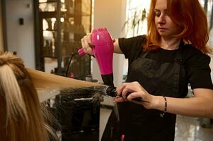 een mooi roodharig kapper gebruik makend van een ronde kam en een haar- droger rechtzetten blond haar- naar een cliënt van een schoonheid salon, het uitvoeren van haar een elegant kapsel foto