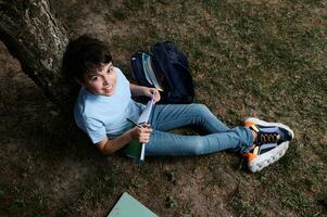 overhead visie van een charmant spaans tiener- school- jongen glimlachen op zoek Bij camera, schrijven Aan schrijfboeken zittend Aan gras foto