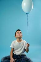 schooljongen jongen zit Aan een blauw achtergrond en looks Bij een blauw ballon. gelukkig kinderjaren concept. vieren kinderen dag foto