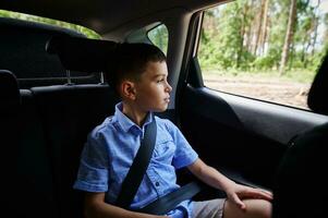 vastgemaakt jongen zittend in de veiligheid auto booster stoel en op zoek uit de venster gedurende reizen door auto foto