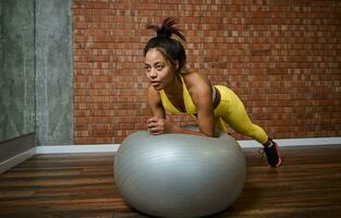 vastbesloten jong Afrikaanse Amerikaans vrouw aan het doen plank Aan een geschiktheid bal Bij Sportschool studio foto