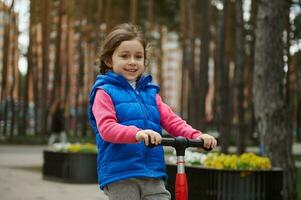 portret van een vrolijk 5 jaar oud Kaukasisch meisje glimlacht een toothy glimlach terwijl rijden een Duwen scooter in een stad park foto