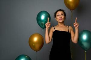 charmant vrouw vervelend een zwart fluweel avond jurk, glimlacht op zoek omhoog en richten met inhoudsopgave vingers Aan een kopiëren ruimte Aan grijs achtergrond met lucht ballonnen. verjaardag, kerstmis, nieuw jaar concept foto