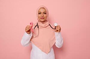 portret van Arabisch moslim vrouw dokter met gedekt hoofd in roze hijaab, poseren Aan gekleurde achtergrond met een roze lint en stethoscoop in handen, ondersteunen kanker patiënten en overlevenden, 1 st oktober foto