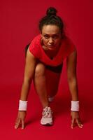 zelfverzekerd vastbesloten atleet vrouw, loper in begin positie klaar naar rennen en sprint, geïsoleerd ovr rood achtergrond met kopiëren advertentie ruimte voor reclame tekst foto