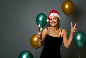 aanbiddelijk vrouw in een de kerstman hoed houdt een fluit met Champagne en shows vrede teken op zoek Bij camera tegen Kerstmis decor met luxe glimmend lucht ballonnen Aan grijs muur achtergrond met kopiëren advertentie ruimte foto