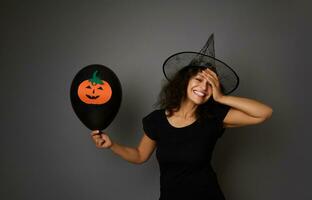 blij lachend vrouw in magie halloween tovenaar hoed looks Bij camera, houdt zwart ballon met vilt gesneden pompoen, glimlacht met mooi toothy glimlach Aan grijs achtergrond. halloween concept, kopiëren ruimte foto